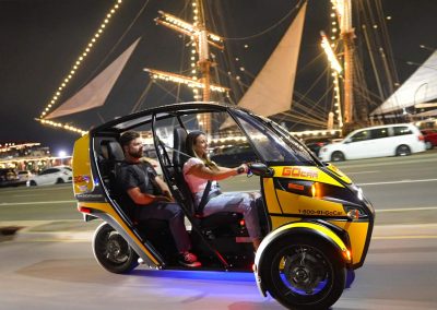 A yellow and black three-wheeled vehicle driving at night with illuminated sails in the background.