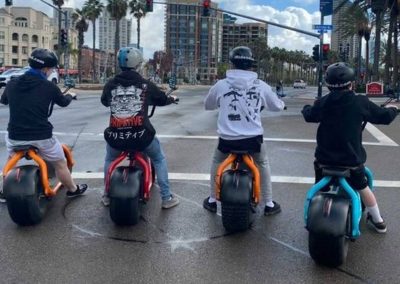 Four people on colorful scooters at a city intersection, waiting at a red light.