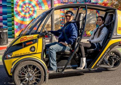 A yellow and black three-wheeled GoCar with two occupants on a colorful street.