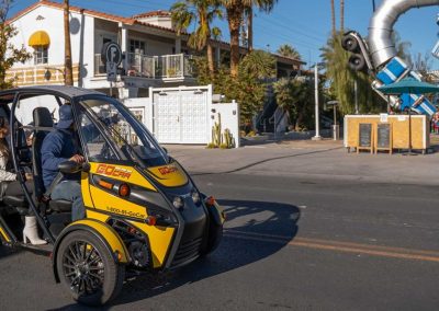 A yellow and black three-wheeled vehicle with two occupants on a sunny street.