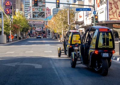 Two electric three-wheeled vehicles driving on a sunny city street with urban surroundings.
